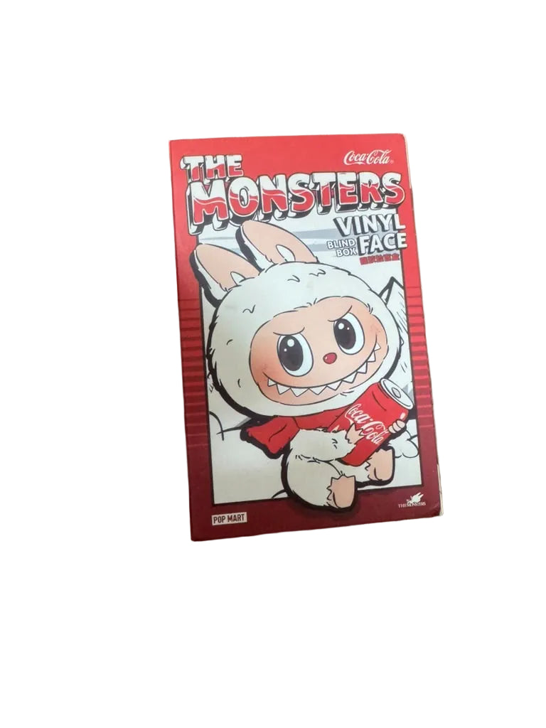 A red box of 'The Monsters Vinyl Face' collectible figures by Coca-Cola, featuring a cartoon character in a white monster suit, placed on a wooden floor.